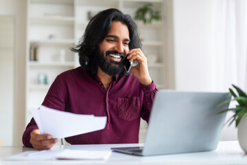 Indian man discussing work on phone while reviewing documents and using laptop - obrazy, fototapety, plakaty