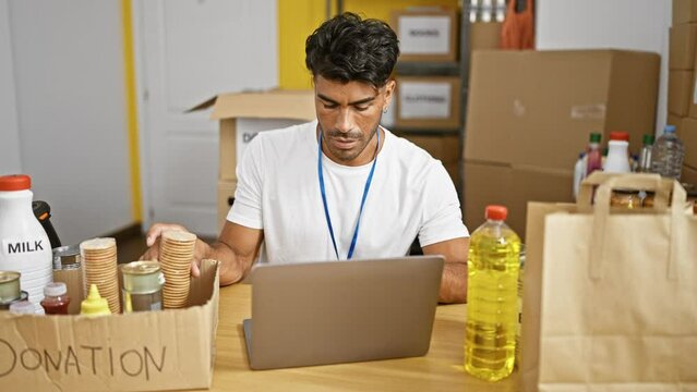 A focused hispanic man volunteers at a donation center, managing food supplies and working on a laptop.