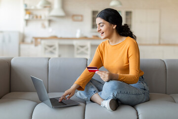 Smiling indian woman sitting on couch, using laptop and holding credit card