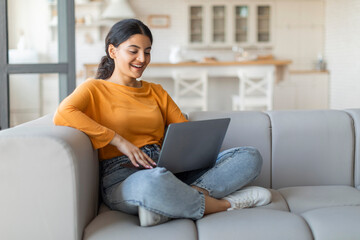 Smiling young indian woman using her laptop while sitting cross-legged on couch