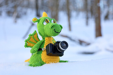 Knitted green dragon with photo camera in a snow on winter forest background. Symbol of Chinese New...