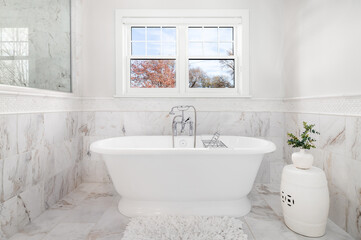 Fototapeta na wymiar A freestanding bathtub with a chrome faucet surrounded by marble tiles on the floor and walls. No brands or labels.