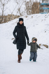 Fototapeta na wymiar Happy mother and son, a beautiful brunette woman and a smiling boy, walk along a snowy road outside in the forest in winter, holding hands. Family photo, vacation concept.