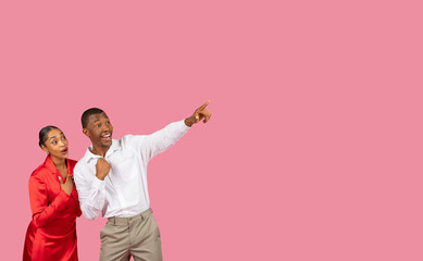 Excited black couple pointing away at free space, wearing elegant attires, pink backdrop