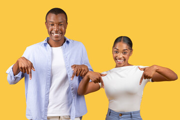 Joyful black couple pointing down, prideful smiles, yellow background