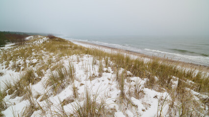 Morze Bałtyckie zimą. Śnieg pokrywa wydmy i rudawozłotą trawę. Mglisty horyzont. Długi czas naświetlania. Rozmyte fale, wyraziste kamyki.