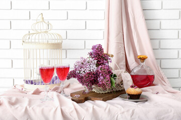 Basket with lilacs and glasses of wine on white table near white brick wall