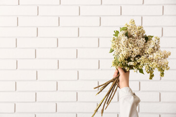 Female hand with bouquet of lilac flowers on white brick wall background