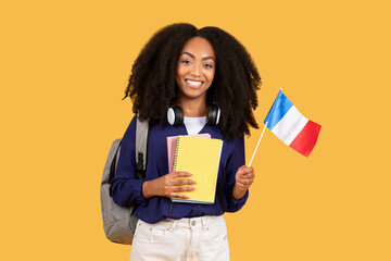 Happy black student with backpack and copybooks, holding flag of France, posing on yellow background