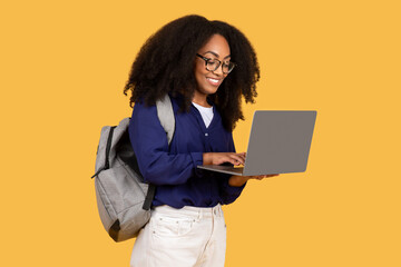 Excited black lady student in glasses with backpack, using laptop and smiling, against yellow background