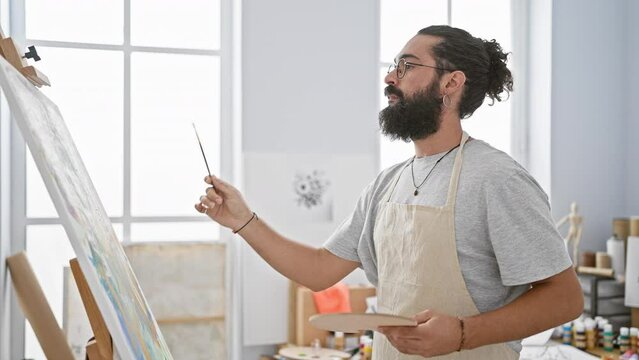 A bearded hispanic man with glasses, painting joyfully in a bright art studio, wearing a casual outfit and an apron.