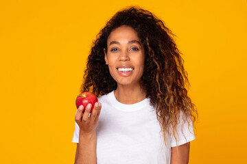 Cheerful black young lady holding fresh red apple, yellow backdrop