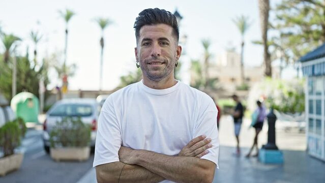 Smiling young hispanic man radiates confidence while casually standing with crossed arms - a joyful expression on sunny city street