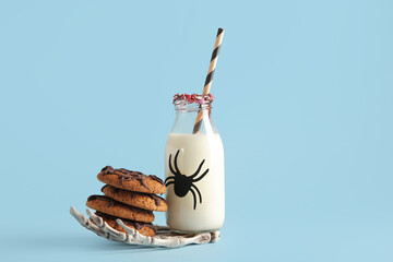 Bottle of milk, skeleton hand and tasty cookies for Halloween on blue background