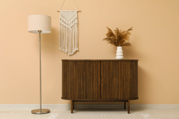 Modern brown chest of drawers, floor lamp and vase with pampas grass near beige wall in room