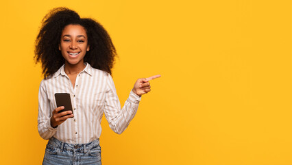 Happy black lady with phone pointing sideways at free space on yellow