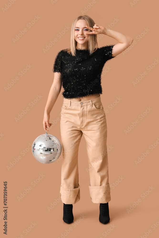 Wall mural Beautiful young woman with disco ball showing victory gesture on brown background