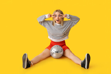 Beautiful young woman with disco ball showing victory gesture on yellow background