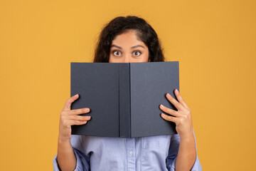 Surprised young woman with curly hair peeking over an open dark book