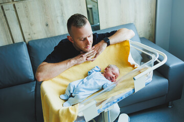 A cute young father is sitting over the crib of his newborn son. A father hugs a newborn baby.