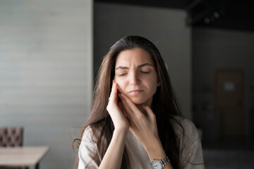 A woman is captured clutching her cheek and closing her eyes in discomfort, indicative of a painful toothache, while in a contemporary indoor environment.