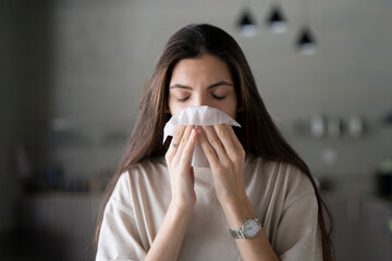 A young woman is captured in the act of blowing her nose with a tissue while seated at a wooden table inside a well-lit room.