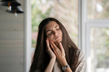 A woman is captured clutching her cheek and closing her eyes in discomfort, indicative of a painful toothache, while in a contemporary indoor environment.