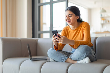 Happy indian woman using smartphone, sitting on sofa with laptop beside her