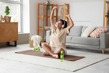 Sporty young African-American woman with headphones sitting on mat in living room