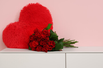 Beautiful bouquet of roses with heart-shaped pillow on commode near pink wall. Valentine's Day...