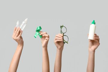 Female hands with bottles of solution, eye drops, green ribbon and eyeglasses on grey background....