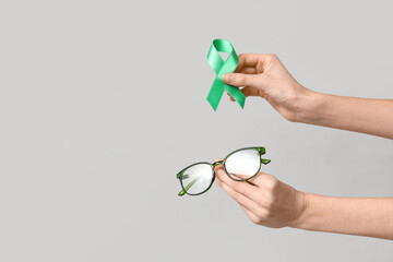 Female hands with green ribbon and eyeglasses on grey background. Glaucoma awareness concept