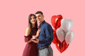 Young engaged couple with wedding ring and heart-shaped balloons on pink background. Valentine's...