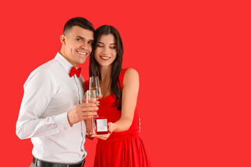 Young engaged couple with wedding ring and glasses of champagne on red background. Valentine's Day celebration