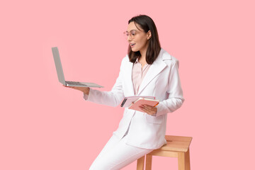 Young businesswoman with laptop sitting on chair against pink background