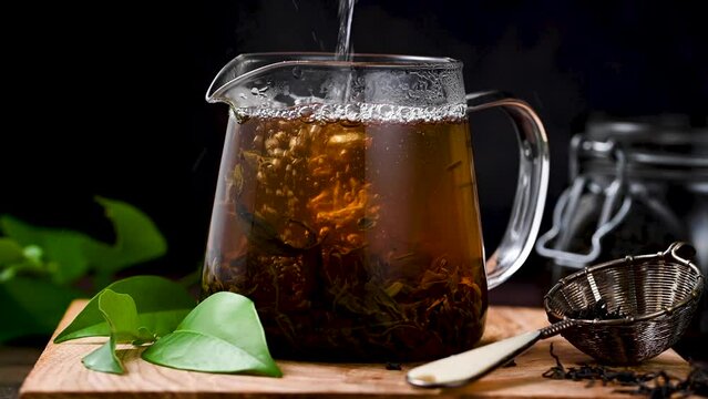 Pouring fresh, hot tea in a glass cup and dry loose leaves, dark moody background