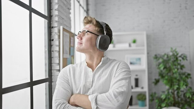 Serious young caucasian man, a business worker listening to his favorite music, while looking out the office window, deep in concentration with arms crossed in indoor professional environment.