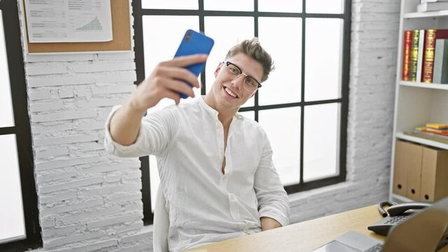 Handsome young caucasian man, confident business worker, making an enjoyable selfie with his smartphone, beaming with a smile at his elegant office desk.