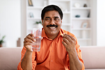 Middle age man taking pill drinking water sitting on sofa