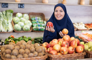 Delighted young Muslim woman purchaser choosing red apples at the counter in grocery store