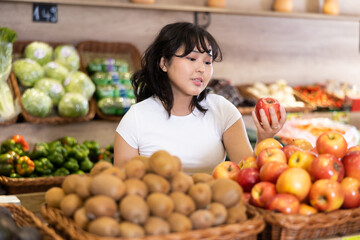 Pleased young woman customer taking ripe apples at the counter in large greengrocery