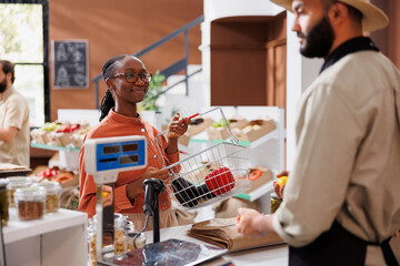 Eco conscious black woman asking about organic, locally grown fruits and vegetables at sustainable supermarket. Male vendor with a hat helping his customer at the checkout counter.