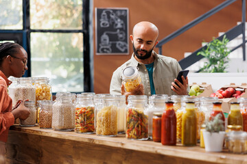 Arab man on his cellphone, surrounded by eco-friendly products and reusable jars. Young middle eastern male customer using his smartphone to compare products and prices in local market.