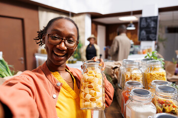 POV of a black woman wearing glasses, holding a glass container filled with pasta in a bio-food supermarket. Online marketing, organic items, vlogging, digital camera, customer filming. - obrazy, fototapety, plakaty