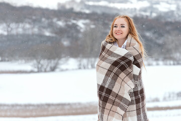 Cute red-haired girl is wrapped in warm knitted blanket and stands outside during the snowfall enjoying the cold winter air.