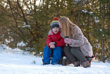 Fototapeta na wymiar Mother and son sitting on a sled in the winter forest chatting and drinking tea from a thermos