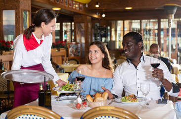 Hospitable young beautiful waitress, bringing dishes to couple of guests at restaurant