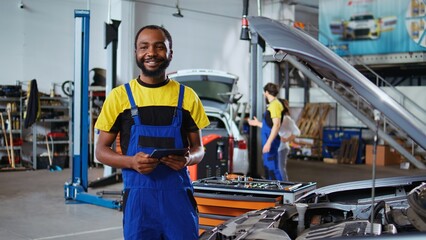 Portrait of smiling BIPOC mechanic in garage using tablet to follow checklist while doing...