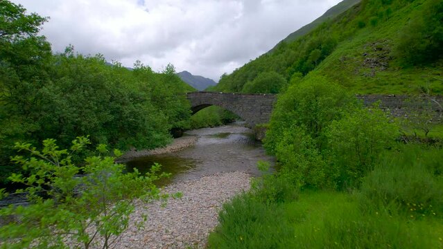 AERIAL: Charming stone bridge over River Shiel which flows amidst lush greenery and meanders between green grassy mountains. Picturesque landscape of the Scottish Highlands with a touch of the past.