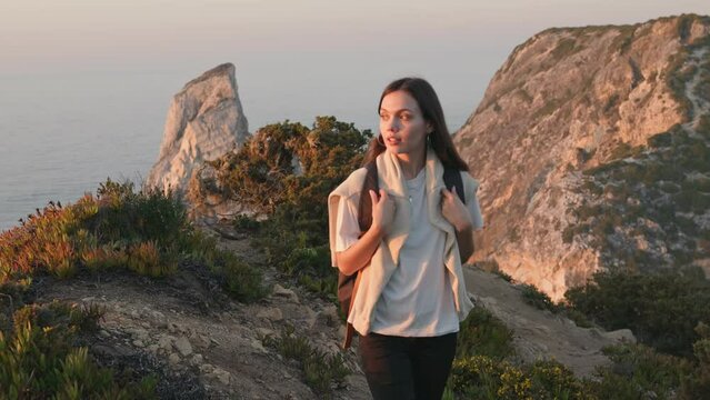 A beautiful young woman enjoys a scenic hike, walking along a cliff edge with a breathtaking sunset over the ocean